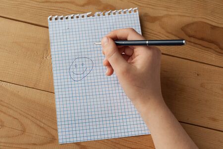 A child is drawing smiley face with the ballpoint pen on the blank squared paper. The ballpoint pen and the blank squared paper, which are from notebook, are on the wooden deskの写真素材