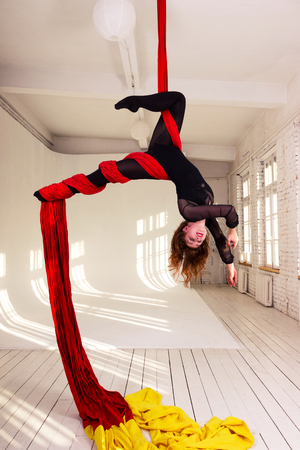 Beautiful aerialist girl doing acrobatic and flexible tricks on red aerial silks (tissues) on white brick and white wall on backgroundの写真素材