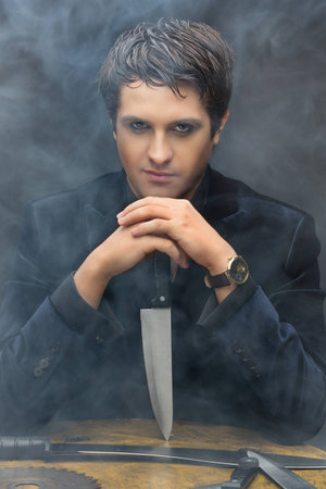 Handsome young man dressed in black suit sitting behind wooden table and holding big knife black smoky backgroundの写真素材