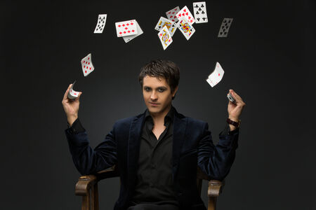 Handsome young man dressed in black suit juggling playing cards over black backgroundの写真素材