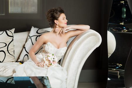 Beautiful young woman in wedding gown sitting on white sofa in interiorの写真素材