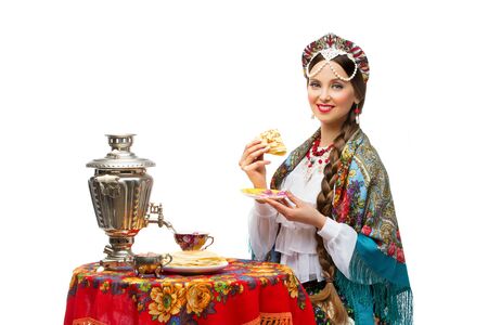 Beautiful young woman in traditional Russian costume sitting in front of table with pancake in her hand. Isolatedの写真素材