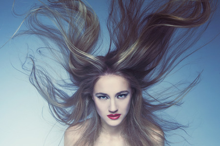 Beautiful young woman with bright make-up and long flying blond hair over blue backgroundの写真素材