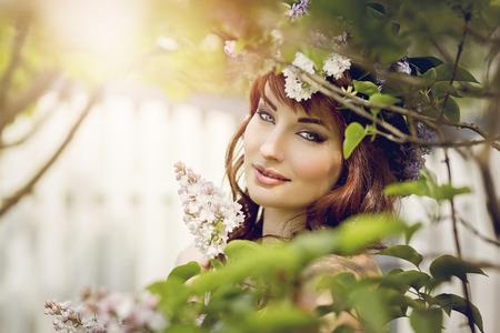 Close-up portrait of beautiful young woman with red hair and lilac on head standing behind tree branches. Outdoor shot.の写真素材