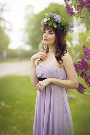 Beautiful young woman in long violet dress with lilac wreath on head standing near tree. Outdoor shot.の写真素材