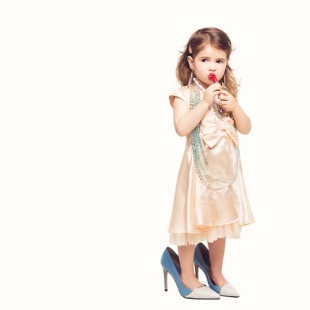 Beautiful small girl in dress and mother's high heels applying red lipstick. Isolated over white background. Copy space.の写真素材