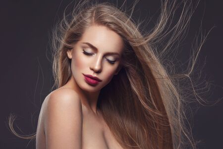 Beautiful young woman with natural makeup and blowing long blond healthy hair. Over dark grey background. Copy space.の写真素材