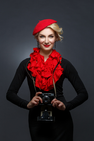 Beautiful young woman in red beret holding retro photo camera. Happy expression. Red lips. Over black background. Copy space.の写真素材