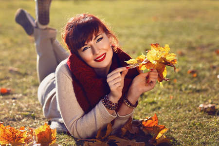 Beautiful young woman lying on ground and holding fall yellow maple leaves in hands. Outside shot....