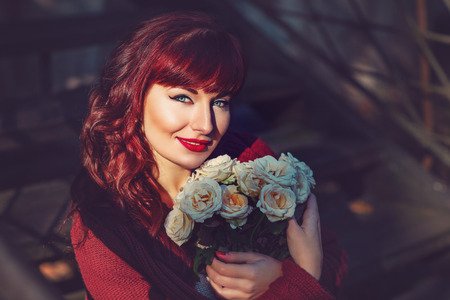 Beautiful young woman in wool red scarf sitting on old house stairs holding flowers. Early fall. Outside shot. Ambient light. Closeup portrait. Copy space.の写真素材