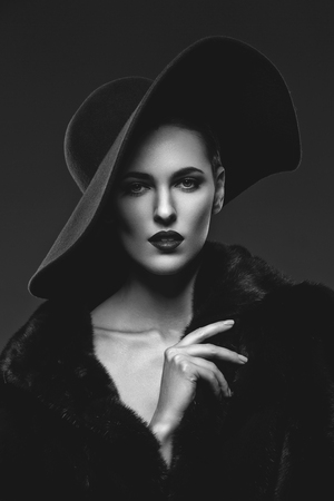 Beautiful young woman with glamorous make-up in luxurious fur coat and fashion hat on black background. Copy space.の写真素材