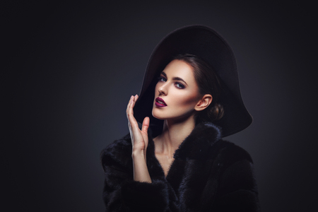 Beautiful young woman with glamorous make-up in luxurious fur coat on naked body and fashion hat on black background. Copy space.の写真素材