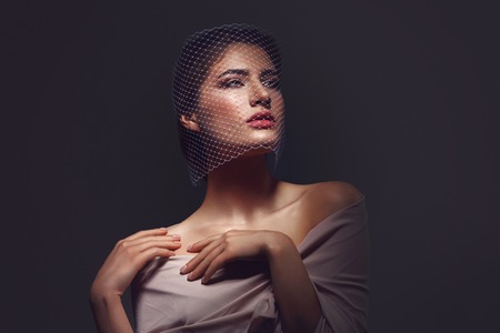 Portret of beautiful young woman with natural makeup. Face covered in veil net. Hands with beige nude manicure. Studio shot on black background. Copy space.の写真素材