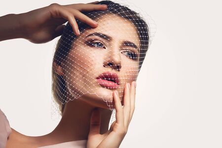 Portret of beautiful young woman with natural makeup. Face covered in veil net. Hands with beige nude manicure. Studio shot. Isolated on white background. Copy space.の写真素材