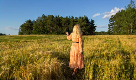 Young woman standing in a wheat field with sunrise on the background. Blonde in a pink dress.
