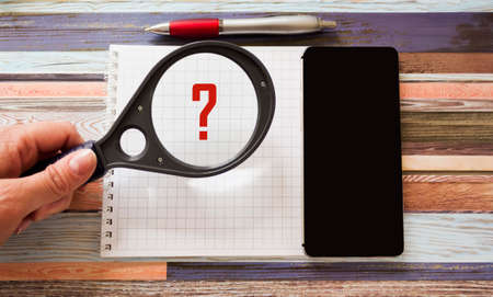 On a striped background lies a notebook with a telephone. A magnifying glass with a question mark is held by a woman's hand.の写真素材