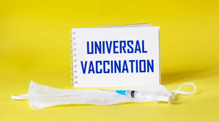 UNIVERSAL VACCINATION is written on a notebook, next to an injection and a medical mask on a yellow background.の写真素材