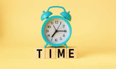 On a light yellow background, wooden cubes with the word TIME and with a blue alarm clock from aboveの写真素材