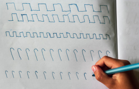 Children's hands with a blue pen on the table with a sheet of paper writes squiggles. View from...