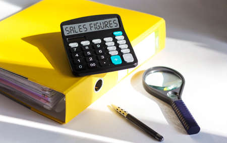 Sales numbers - text on card next to document folder, magnifier, calculator and pen on white...