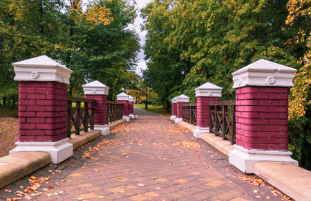 Red brick columns on the fence of a bridge in an autumn park with yellowed trees. Beautiful autumn...