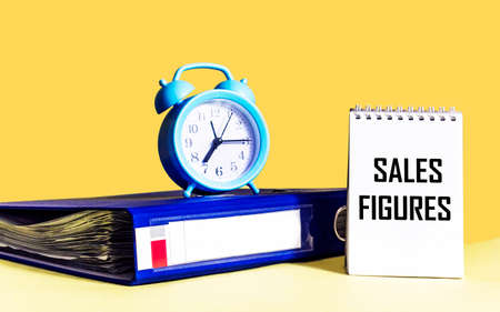 Sales Figures - text on a notebook next to the folder and the clock.