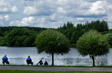 Two men dressed in blue jackets are fishing together with a fishing rod, sitting on the shore of a lake.の写真素材