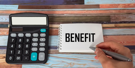A woman's hand writes BENEFITS on a notepad, next to a calculator on a beautiful background.の写真素材