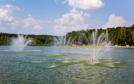 Lake with water cannon and fountain breeze, green leafy trees in the background, sunny summer day with clouds in Europeの写真素材