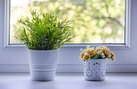 Yellow decorative flowers in a white flower pot and a pot with green leaves on the window