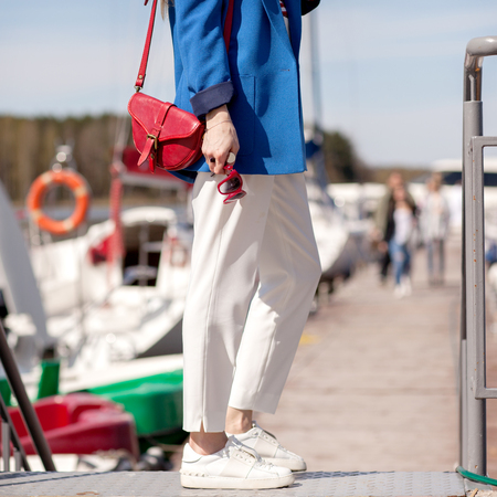 Street, bright style. Young woman in a blue jacket, white sneakers, white trousers, a red handbag, Sunglassesand and a red case for sunglasses. Details. Sguare image photoの写真素材