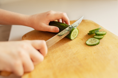 Girl cuts cucumber on a wooden board. Closeup, kitchen, hands, knifeの写真素材