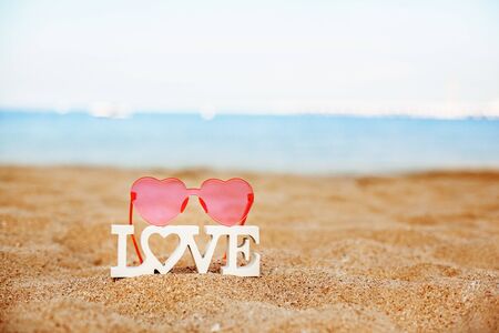 Wooden letters Love and pink heart-shaped glasses on a sandy beach overlooking the blue seaの写真素材