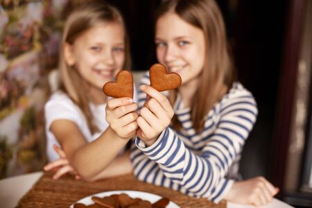 Charming, funny and laughing girls on a blurry background are holding heart-shaped cookies on an outstretched arm, two hearts, close-upの写真素材