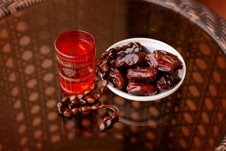 An Oriental drink, an Arabic rosary, and dates on a white plate stand on a glass table. The Islamic...
