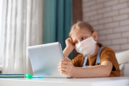A girl in a medical mask is studying at home with a digital tablet laptop and doing homework. Distance learning online educationの写真素材
