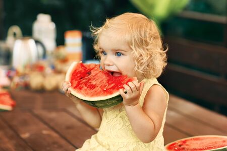 Little blonde girl eating a juicy watermelon in the garden. Children eat fruit on the street. Healthy food for children. Toddler gardening.の写真素材