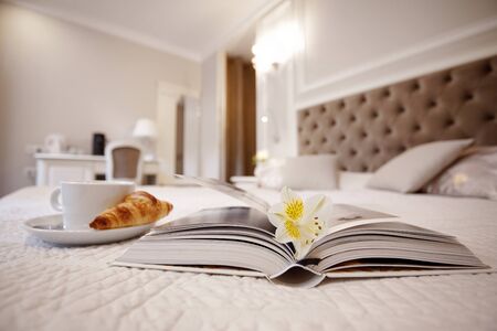 A Cup of coffee, a croissant and an open book lie on the bed in a bright cozy room, a delicate...
