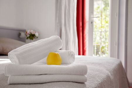 Beautifully and neatly folded white towels lie on the bed in the room against the background of the window. Towels are decorated with a delicious yellow Appleの写真素材
