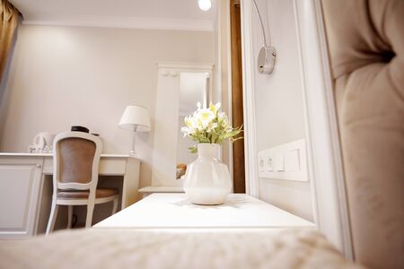 A beautiful vase with white flowers stands near the bed in a bright comfortable room. Time to relax, holiday. Space for textの写真素材