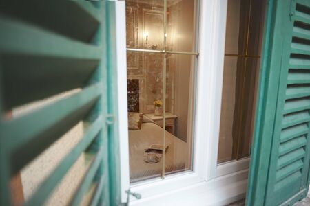 View through the half-open shutters and window on the interior of the bedroom. A Cup of coffee, a...