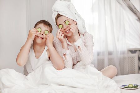 A beautiful young girl and her cheerful brother make a face mask from cucumber slices. The brother and sister are sitting on the bed with cucumbers in their eyes. The concept of beauty and healthの写真素材