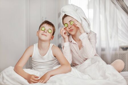 A beautiful young girl and her cheerful brother make a face mask from cucumber slices. The brother and sister are sitting on the bed with cucumbers in their eyes. The concept of beauty and healthの写真素材