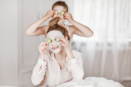 A beautiful young girl and her cheerful brother make a face mask from cucumber slices. The brother and sister are sitting on the bed with cucumbers in their eyes. The concept of beauty and healthの写真素材