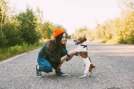 Attractive teen girl playing with her dog while walking in the fresh air. The girl holds the pet by its front paws, crouching next to it. Friendship, care, participationの写真素材