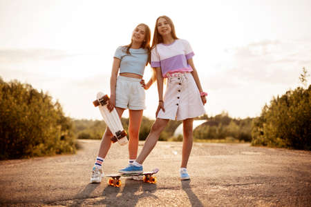 Two blond teenage girls ride skateboards at beautiful sunset.