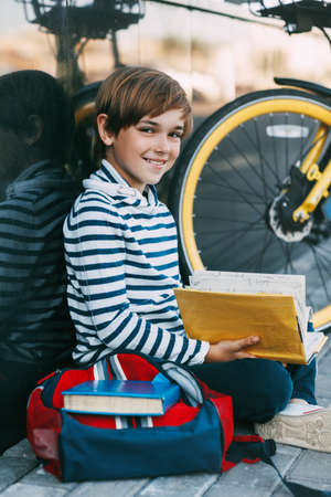 A cheerful boy is sitting on the sidewalk near the school and reading a book. Next to it is a Bicycle and a backpack, portraitの写真素材