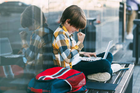 A happy boy in a plaid shirt sits on a bench and holds a laptop in his lap. The boy communicates with friends on the Internet. Social distance, positive emotions. Education, technology,の写真素材