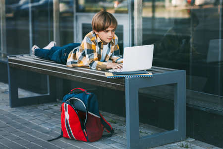 A schoolboy is lying on a wooden bench and working on a laptop, next to a backpack. The boy is preparing for school lessons using the Internet. Social distance. Distance learning, education, technologyの写真素材