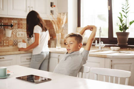 A tired boy pulls himself up after online lessons in the kitchen at home, his sister helps him. Distance learning, online education, home schooling conceptの写真素材